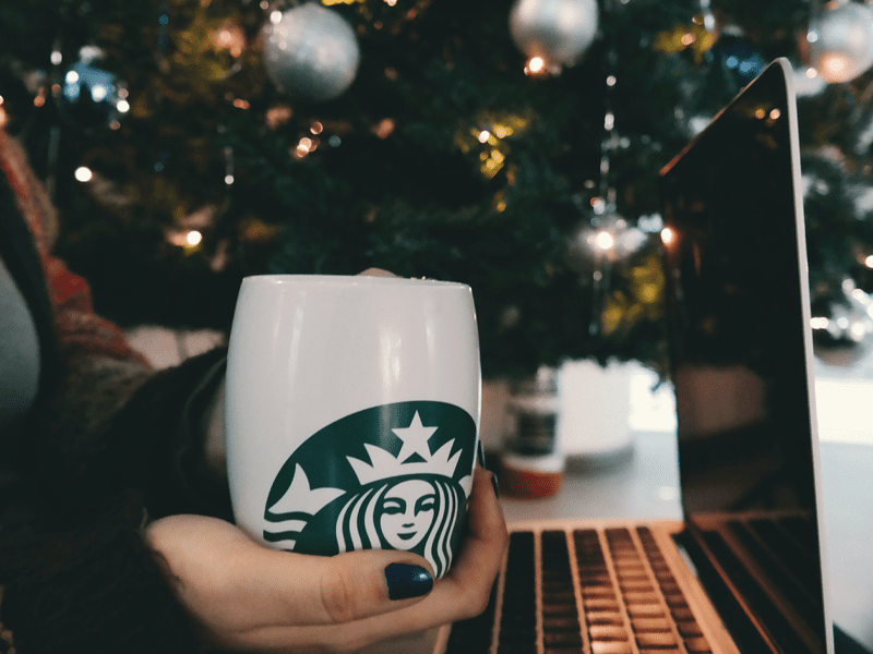 Hand holding cup with laptop on lap next to Christmas tree