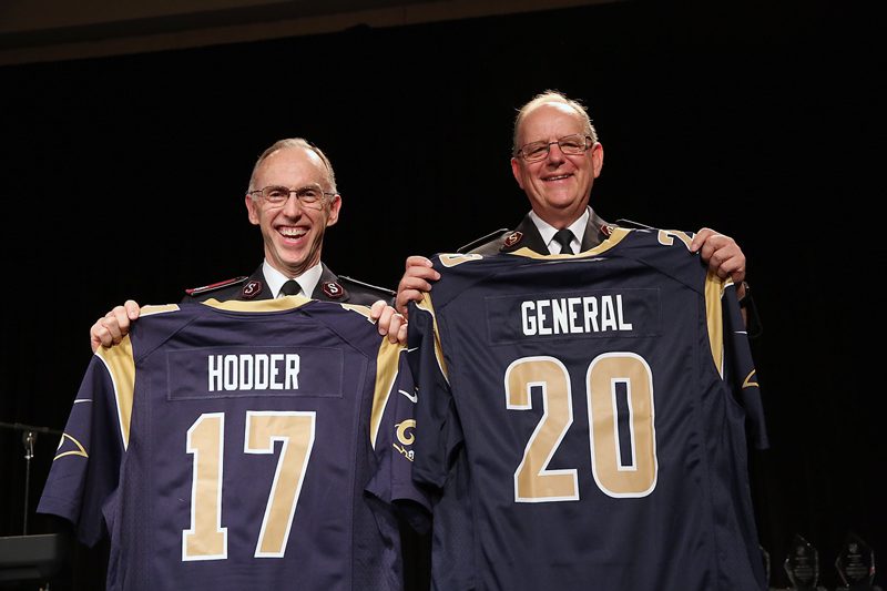 Commissioner Kenneth G. Hodder and General André Cox hold up custom embroidered Los Angeles Rams jerseys from Major Kyle Trimmer at the Men's Ministries breakfast Saturday morning.