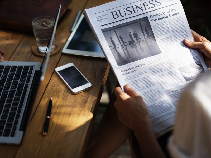 person reading newspaper next to phone and laptop on table 