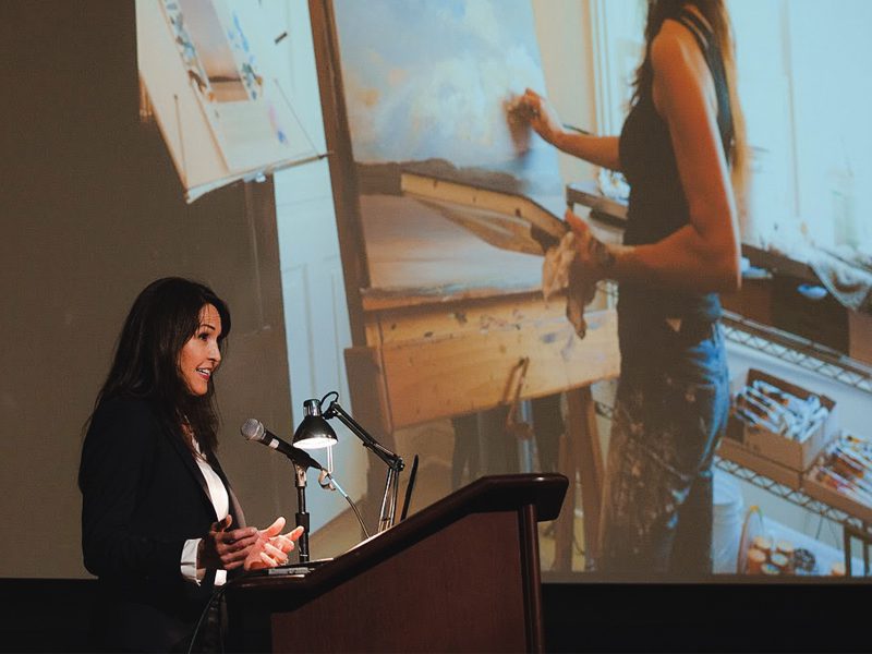 Woman speaking at podium with projection of painter in the background