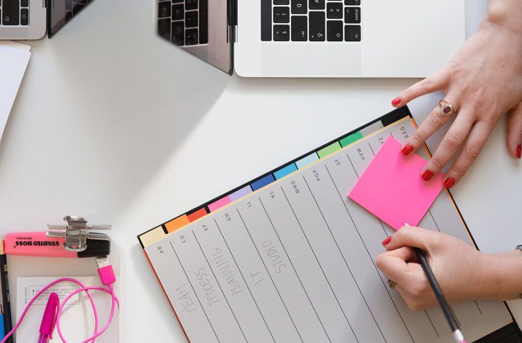 hands writing in personal calendar with post it note next to laptop