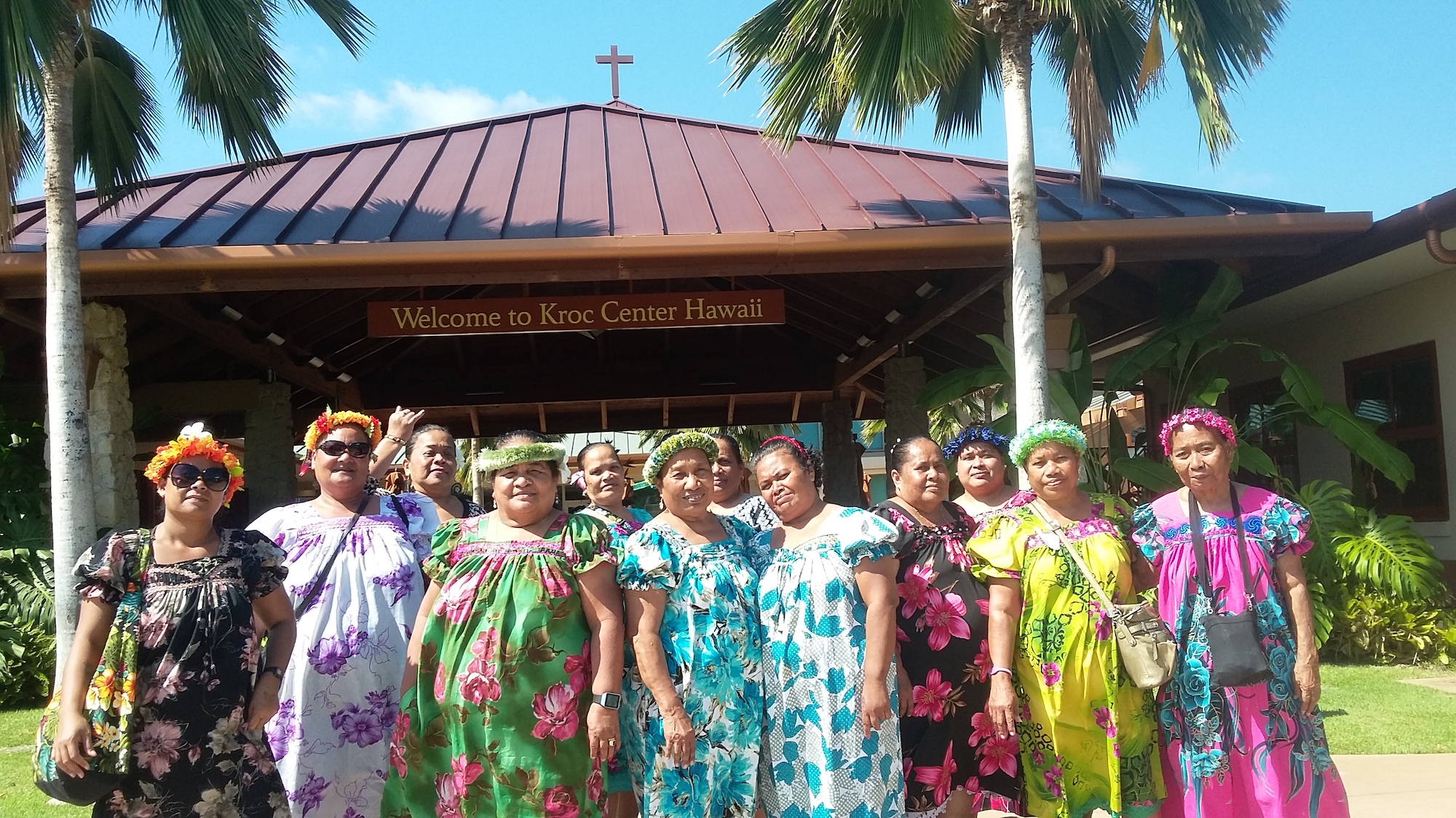 Women smile and stand in front of a building wearing colorful dresses