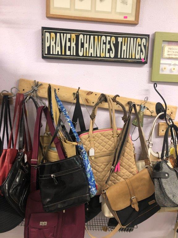 a sign that reads 'prayer changes everything' hangs on the wall in a salvation army thrift store above a rack of handbags
